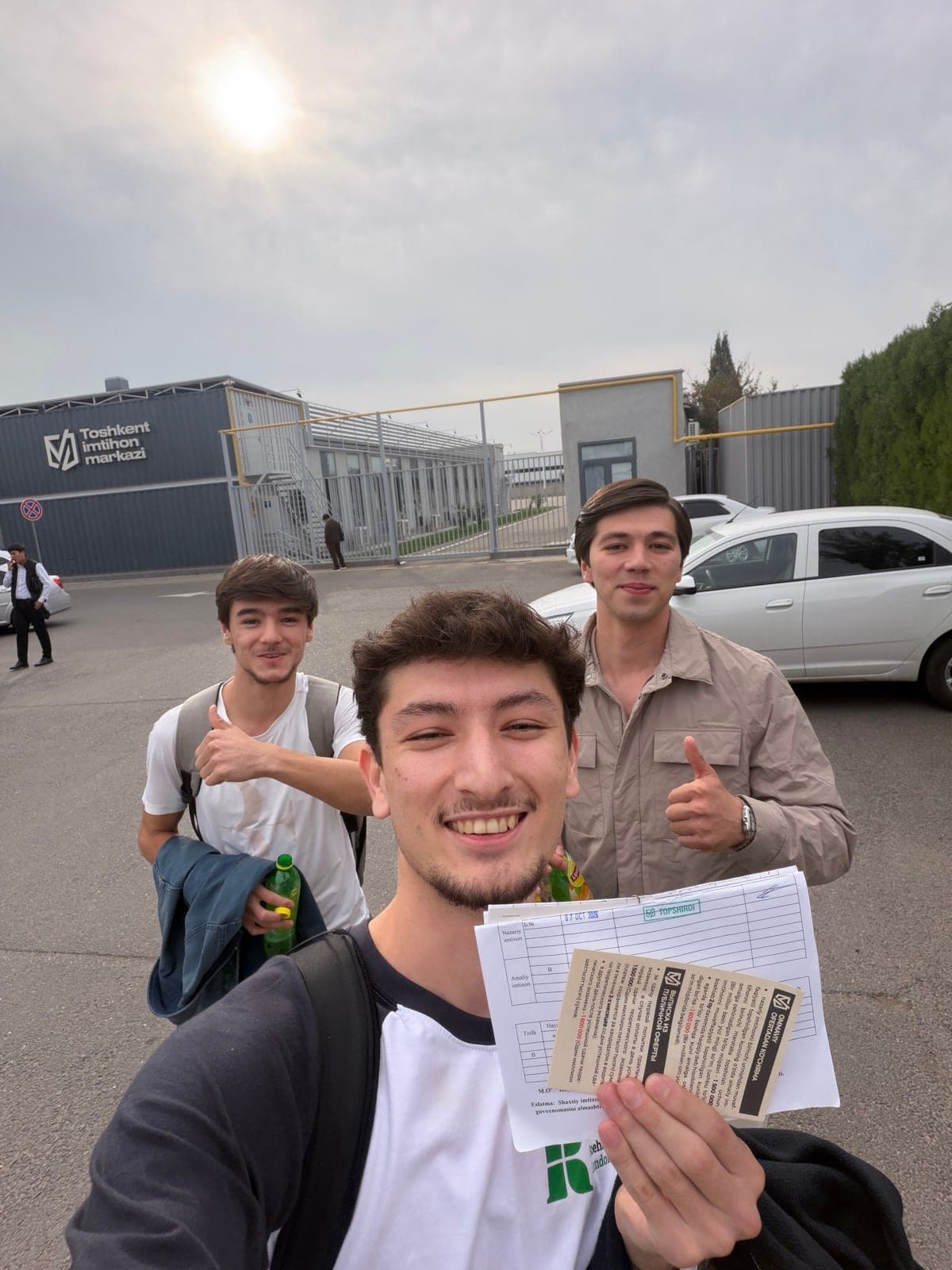 Rahmonberdi (Tommy) Vahabov outside the driving exam centre on 7 October 2026 with two friends, holding his exam sheet stamped TOPSHIRDI